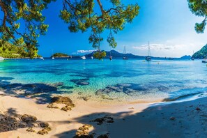 Una playa con agua turquesa cristalina, orilla de arena y barcos a lo lejos.