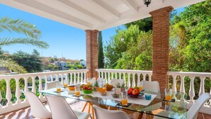 A glass-top table with chairs on a balcony, surrounded by a white balustrade, with a view of greenery and buildings in the background. - Villa Arantxa by Villa Plus (Frigiliana)