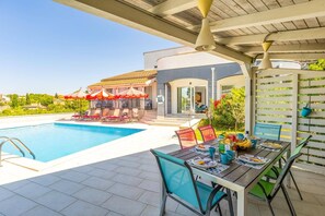 Un espace aménagé au bord de la piscine, avec une table à manger pour deux personnes, un coin salon couvert avec des parasols et une vue dégagée sur la piscine.