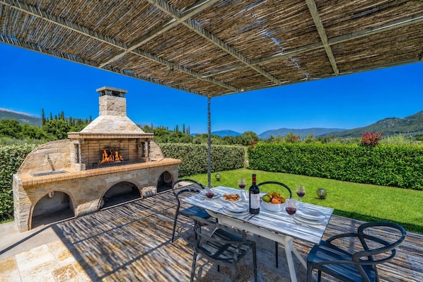 An outdoor barbecue area with a stone oven, a table set for dining, and a view of the surrounding landscape.