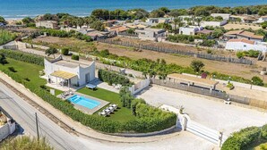 Ett flygbildsbild av ett bostadsområde vid kusten med en swimmingpool, en liten vit byggnad och hus med röda tegeltak.