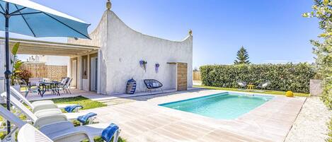 Un área junto a la piscina con sillas de descanso, un porche cubierto y un pequeño edificio de diseño arquitectónico singular.