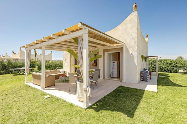 A covered outdoor seating area with a sofa, chairs, and a table, adjacent to a building with a conical roof.