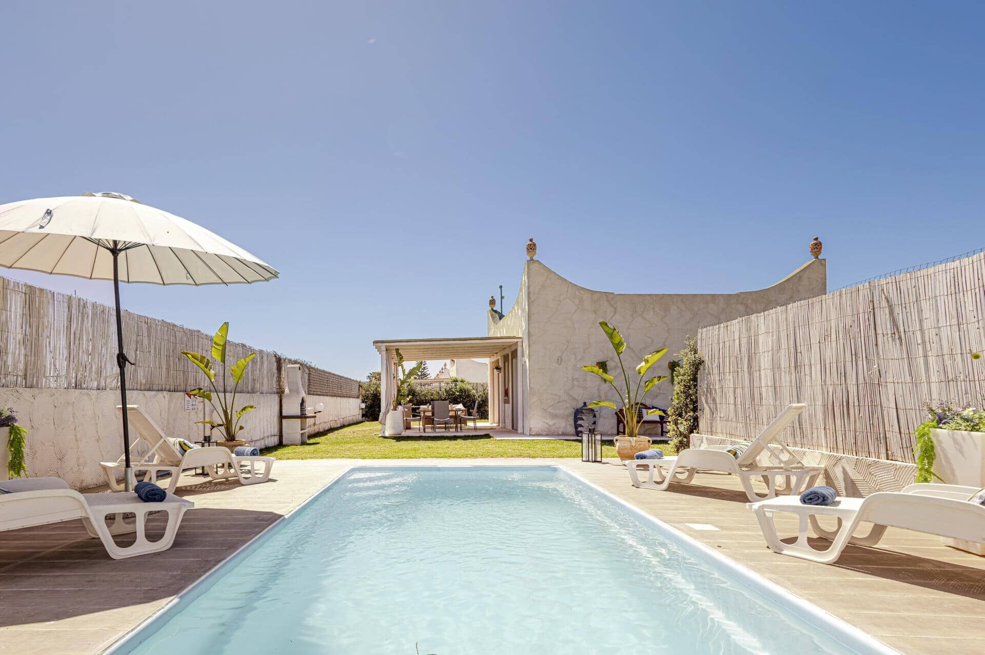 Une piscine entourée d’une clôture en bois et d’un bâtiment à toit incurvé, avec des chaises longues et un parasol à proximité.