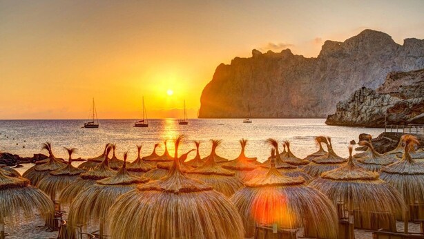 Une plage avec des parasols et des chaises en osier, un coucher de soleil et des bateaux sur l’eau.