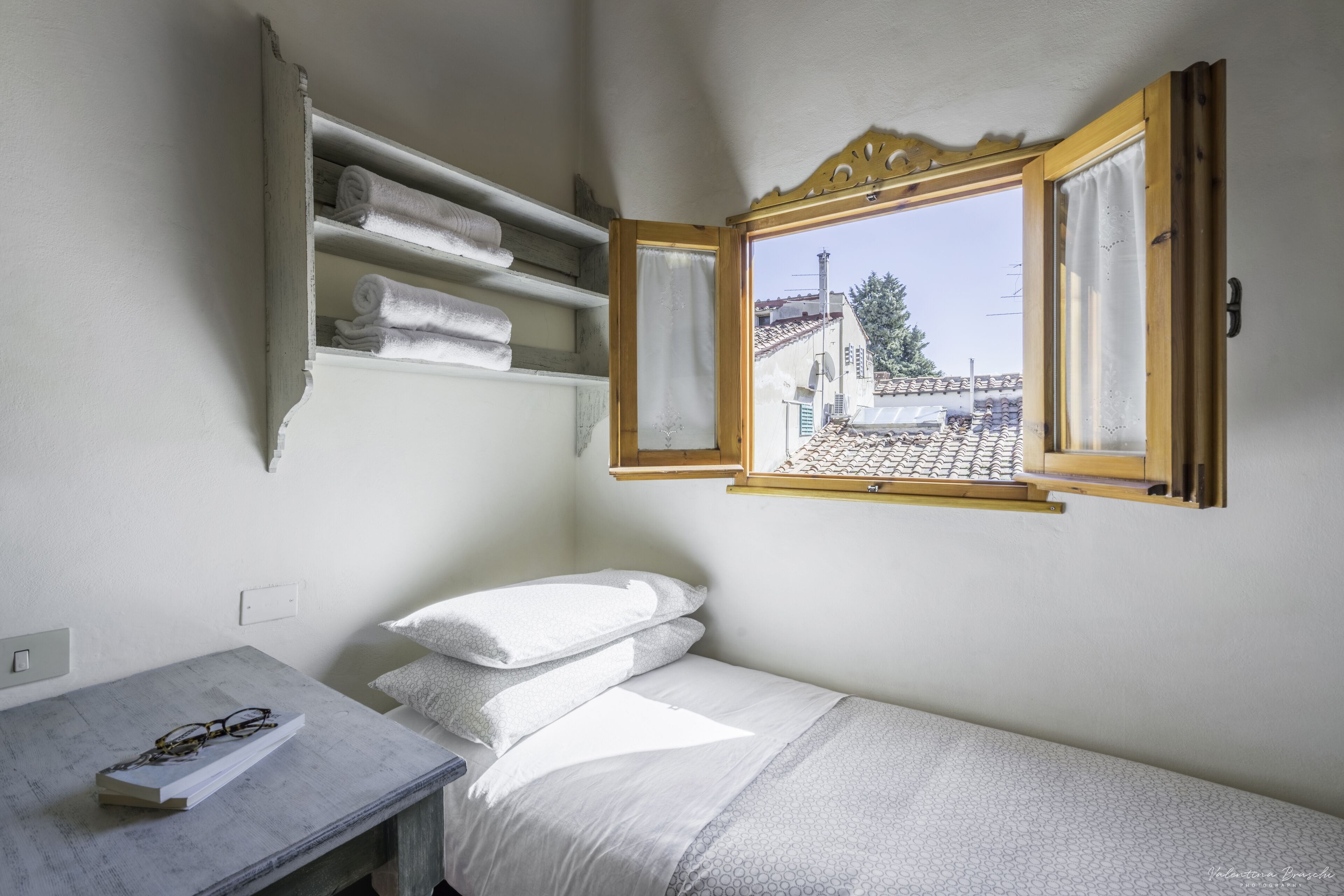 A small, neatly arranged bedroom with a single bed, a bedside table with glasses, and a window with a view of buildings.