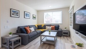 A modern living room with a grey sectional sofa, a coffee table, and a TV on a stand. There are framed pictures on the wall and a window providing natural light.