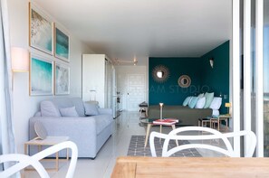 A modern living room with a grey sofa, a dining table with chairs, and a kitchen area visible through an open door.