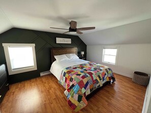 A bedroom with a wooden floor, a bed with a colorful quilt, a ceiling fan, and two windows.