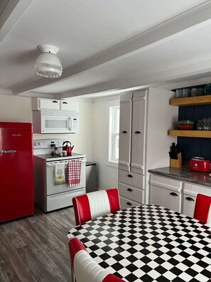 A compact kitchen with a red refrigerator, white microwave, and stove, a checkered table, and a window.