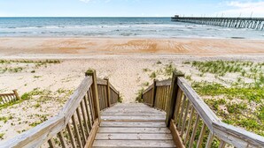 A wooden staircase leading to a sandy beach with a pier in the background.