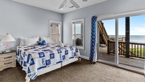 A bedroom with a large bed, a ceiling fan, and a view of a wooden deck and ocean.