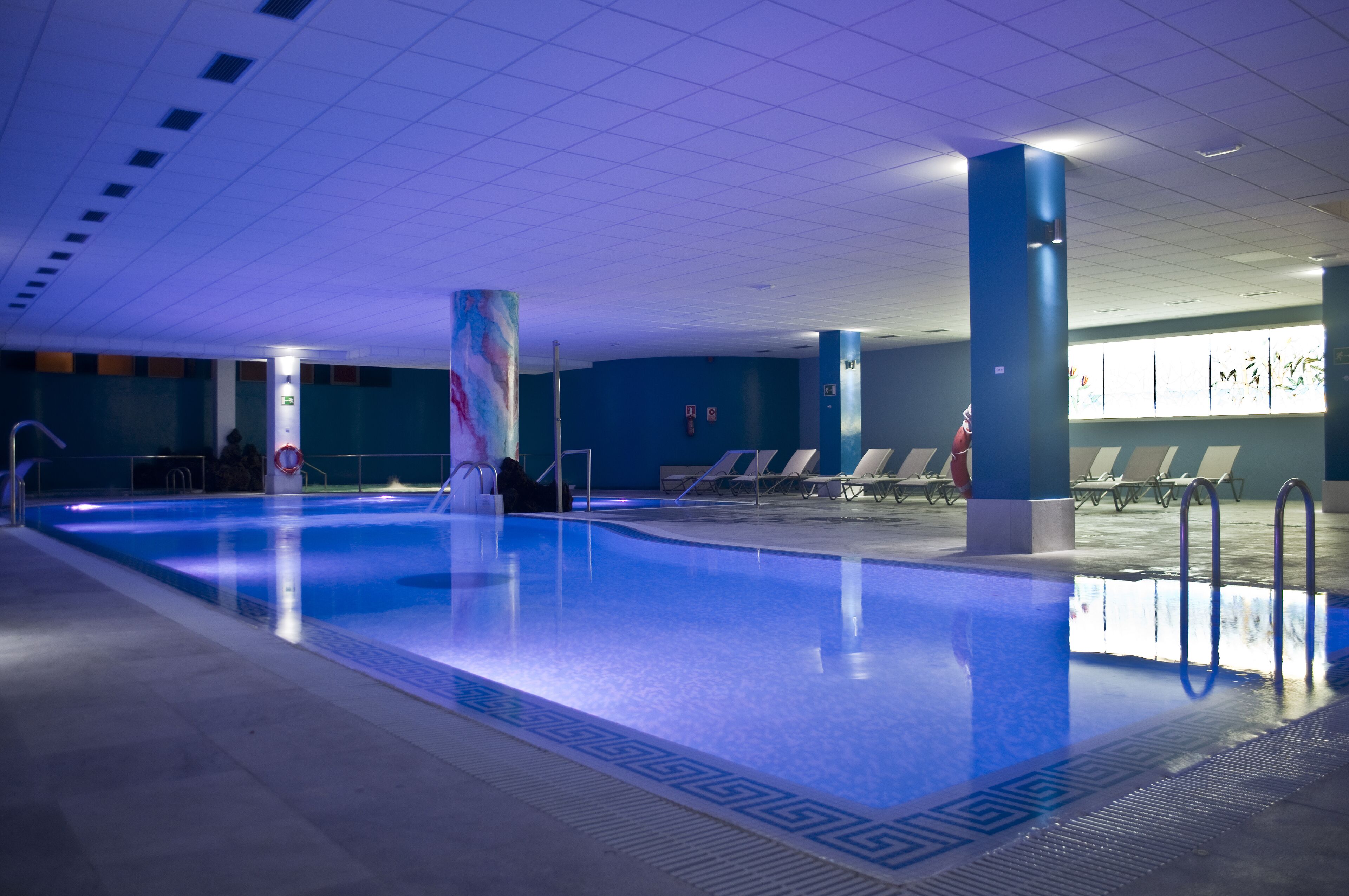 Piscina interna com iluminação azul, cadeiras de descanso e piso revestido de azulejos.