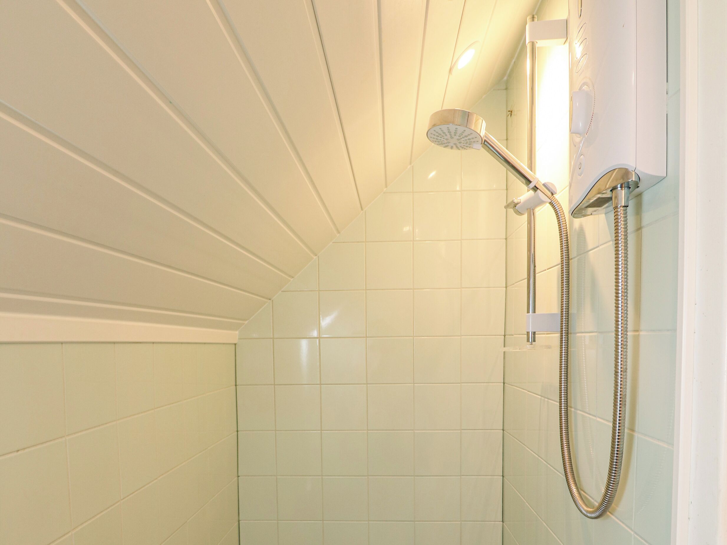 A shower with a handheld showerhead and a flexible hose, tiled walls, and a ceiling with a sloped design.
