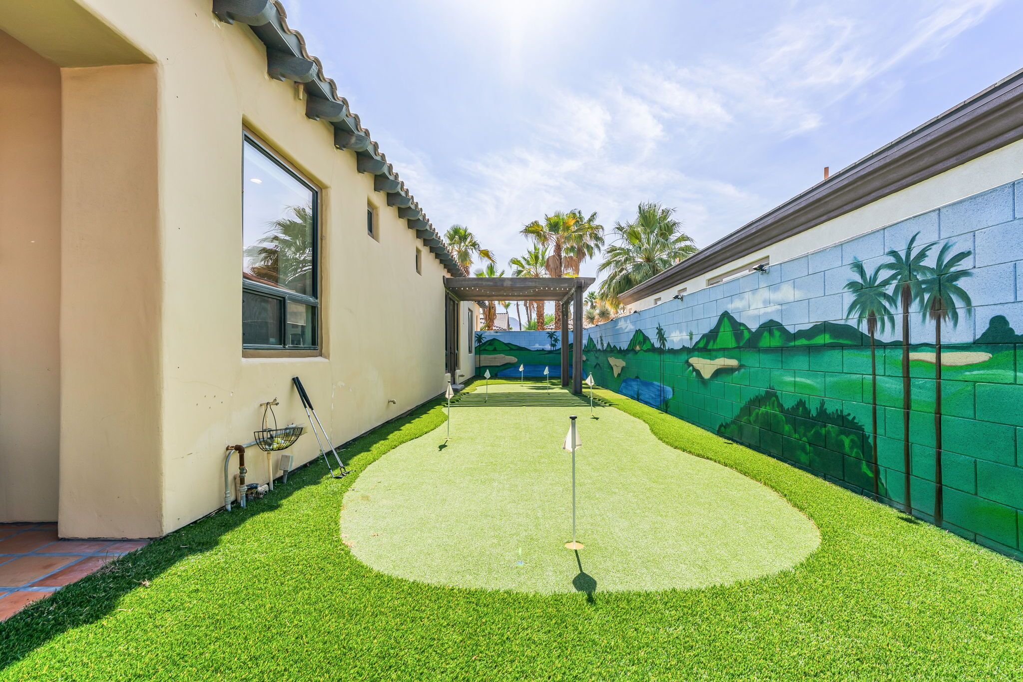 A mini-golf course with a green artificial turf, a pergola, and a mural of a golf course on the adjacent wall.