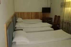 Quarto de hotel estreito com três camas alinhadas na parede, uma televisão montada acima das camas e uma pequena mesa com cadeiras.