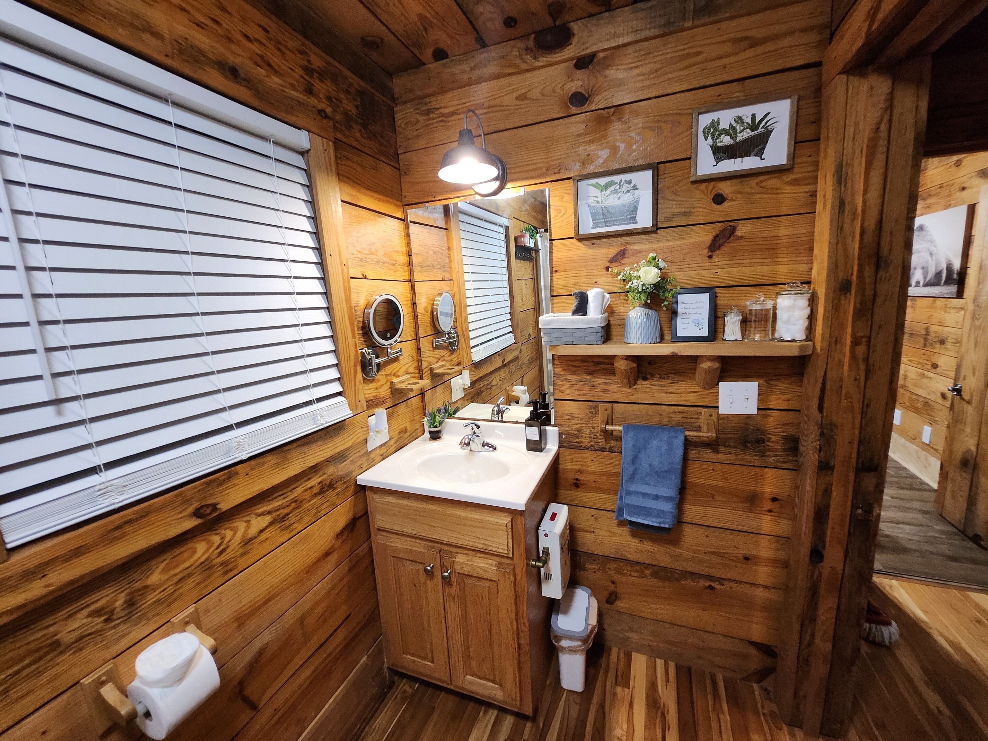 Une salle de bain rustique, aménagée dans une cabane en bois, avec un lavabo, un miroir, des toilettes et des stores aux fenêtres.