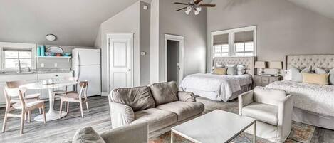 A spacious living area with a high ceiling, two ceiling fans, a dining table with chairs, a sofa, a coffee table, and a bedroom with a bed and nightstands.