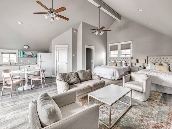 A spacious living area with a high ceiling, two ceiling fans, a dining table with chairs, a sofa, a coffee table, and a large bed with a headboard.