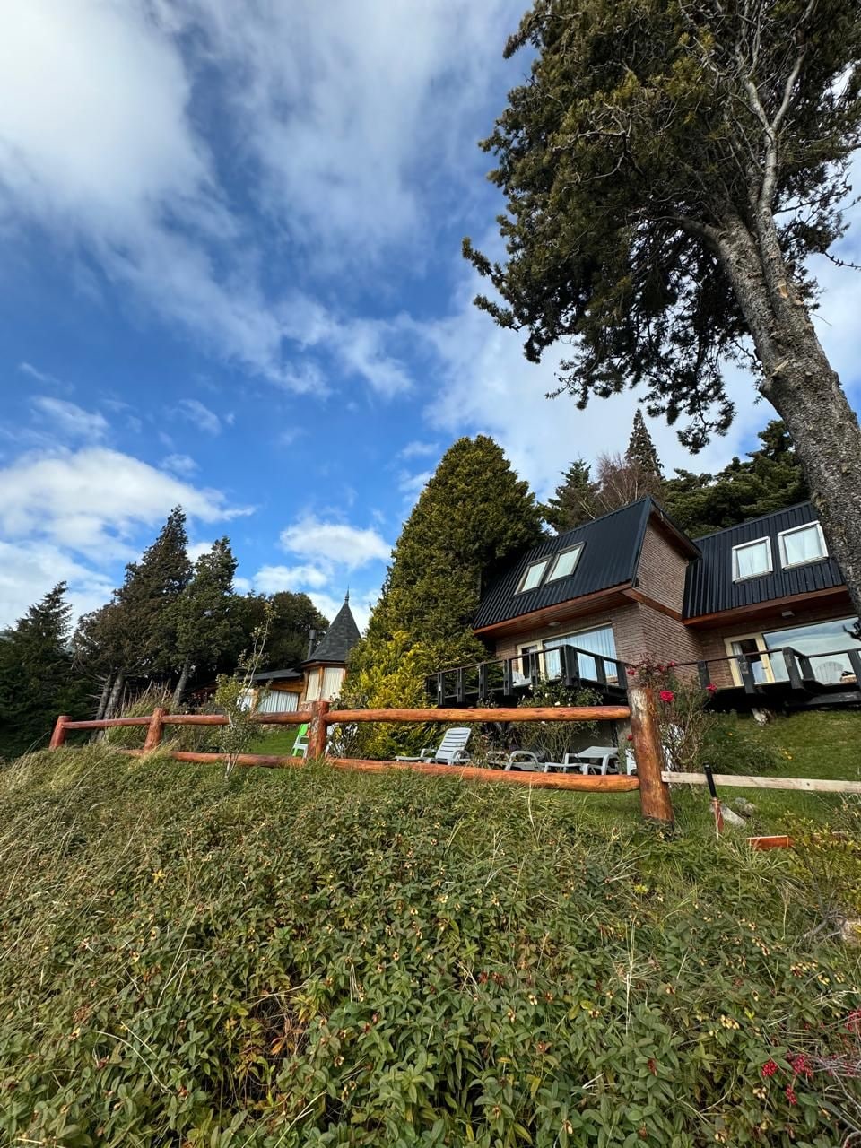 Una casa moderna de dos pisos con un balcón, rodeada de exuberante vegetación y altos árboles.