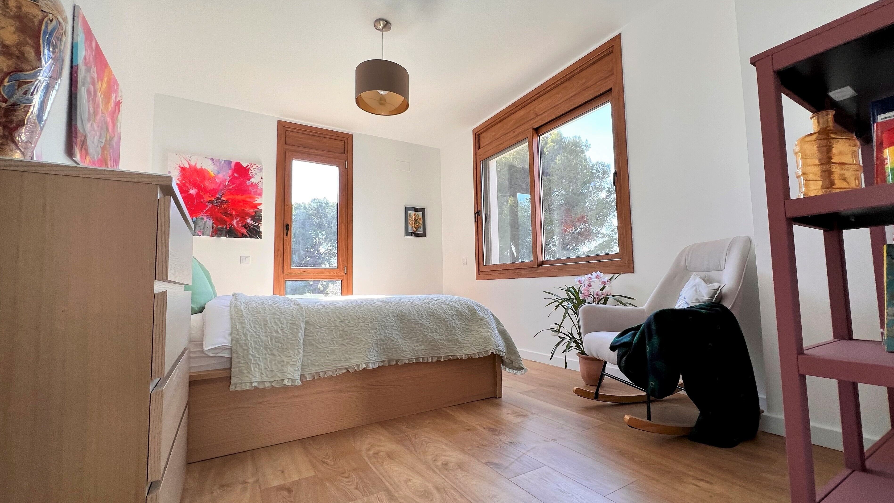 Quarto com cama, uma cadeira, um vaso de planta e uma prateleira com uma garrafa.