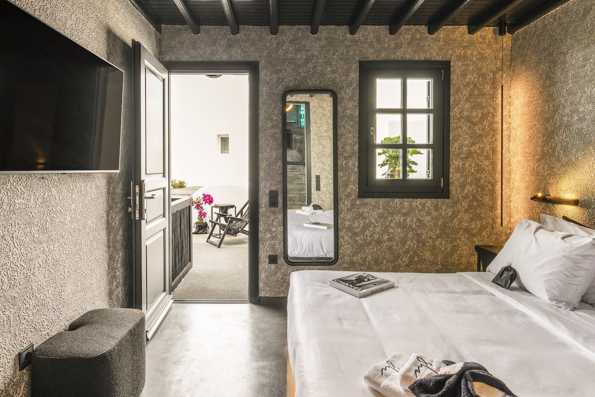 A bedroom with a large bed, a TV, and a mirror. There is a door leading to a balcony with a chair and flowers.