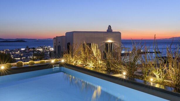 A poolside view with a sunset over the sea, a building with a tower, and a cityscape in the background. - She Mykonos Boutique Hotel (Mykonos)
