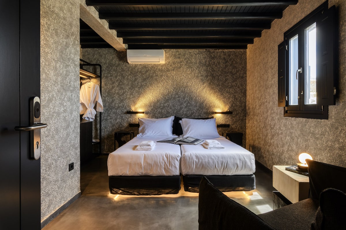 A modern bedroom with two beds, a nightstand, a reading lamp, and a window with curtains.