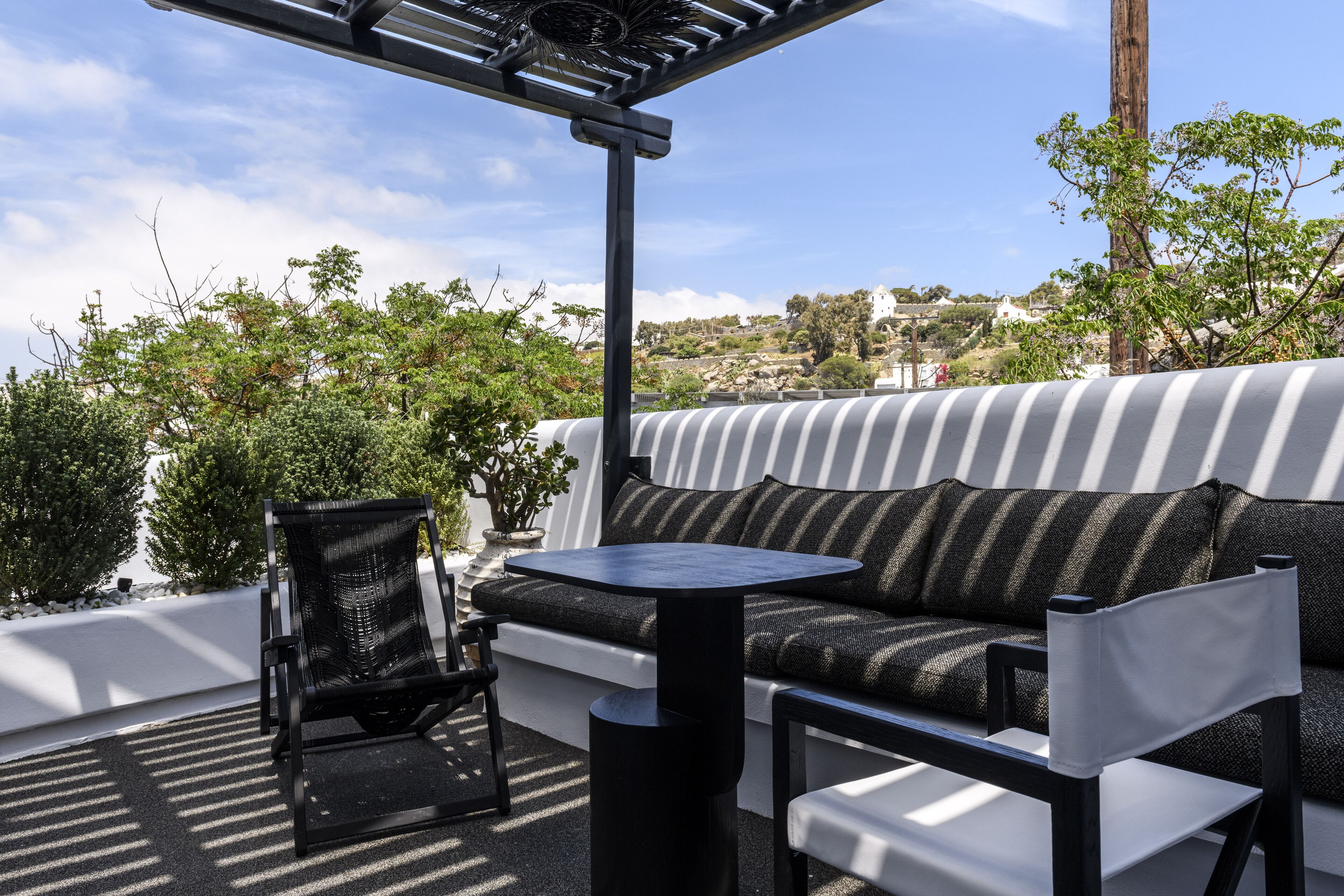 A rooftop terrace with a black metal table, a black metal chair, a black and white striped sofa, and a pergola.