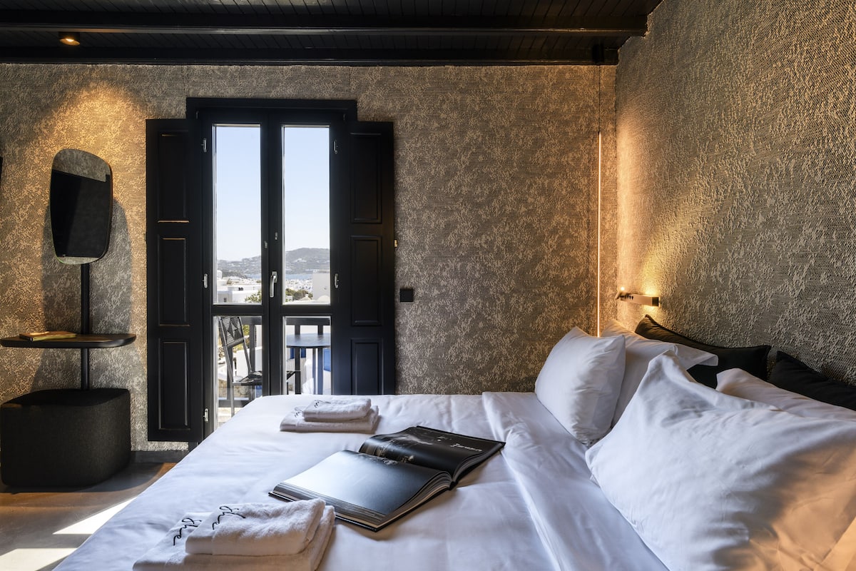 A hotel room with a bed, a book, a towel, a black chair, and a wall-mounted TV.