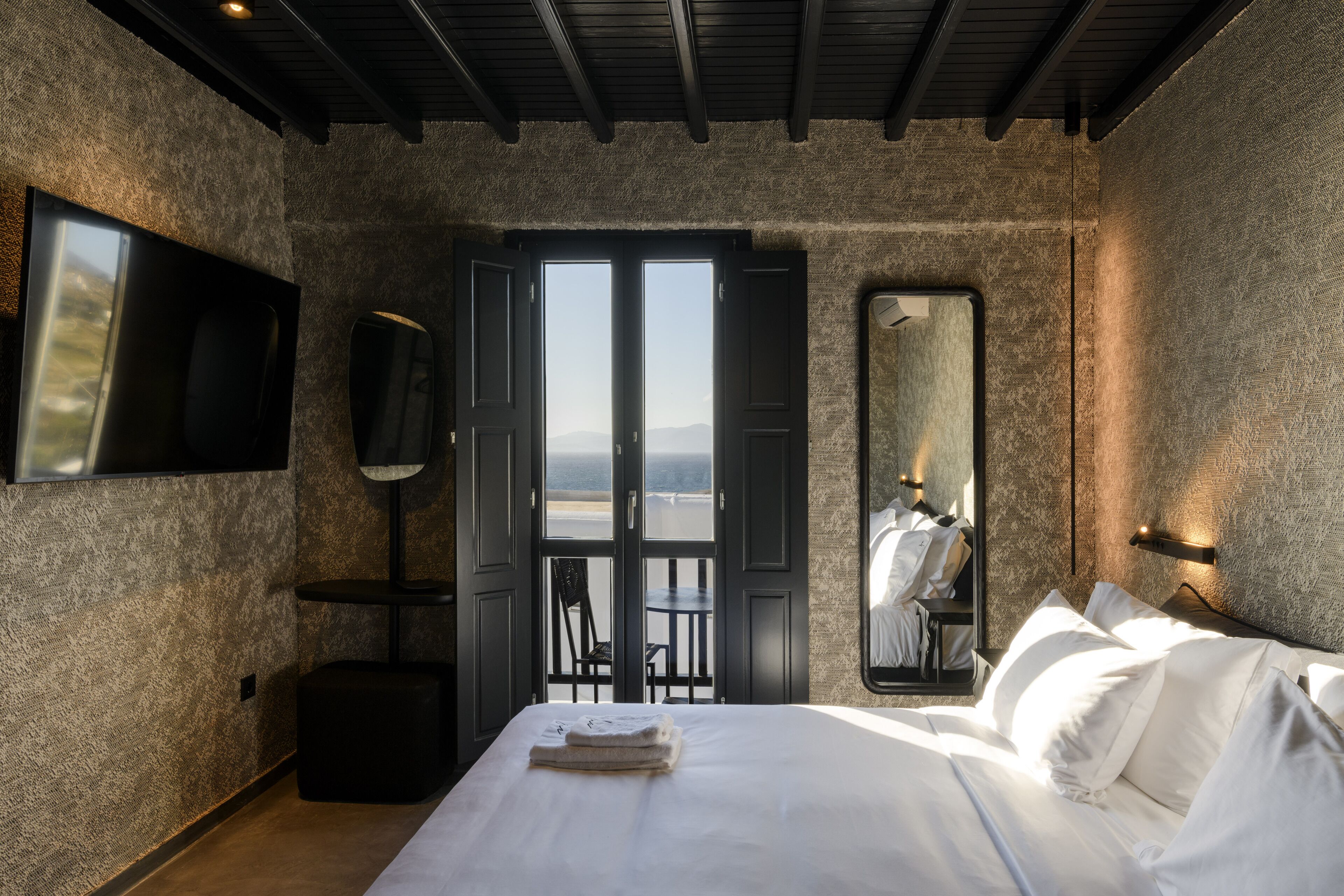 Ein Schlafzimmer mit Bett, Fernseher, großem Spiegel und Blick auf das Meer durch offene Türen.