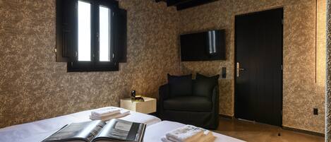 Ein Hotelzimmer mit einem großen Bett, einem Fenster mit Rollläden, einem an der Wand befestigten Flachbildfernseher, einem schwarzen Sessel und einem Holznachttisch mit Lampe.