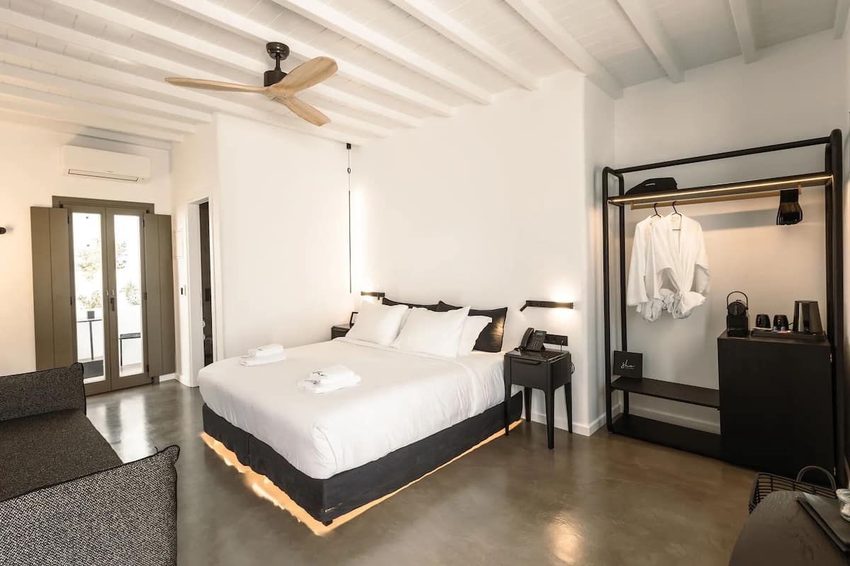 Una habitación de hotel moderna con una cama grande, un ventilador de techo, una mesita de noche con una lámpara, un perchero para bata y un pequeño escritorio con un teléfono y una cafetera.