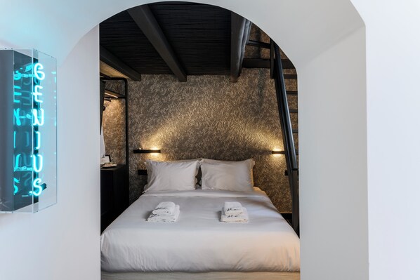 A neatly made bed with white linens and towels, a stone-textured wall, and a neon sign on the left. - She Mykonos - The Residency (Mykonos)
