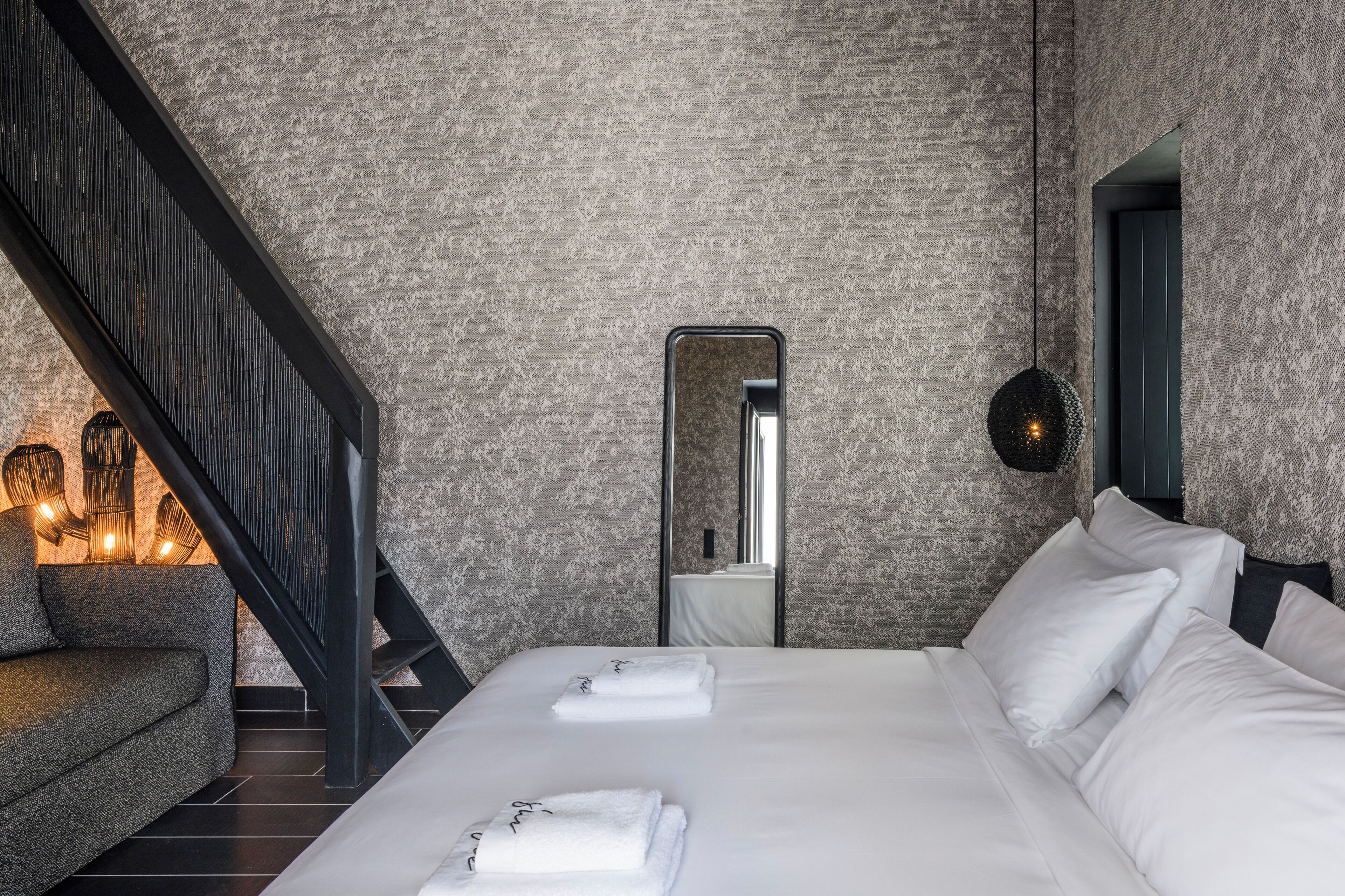 A modern hotel room with a large bed, a sofa, a staircase, and a mirror.