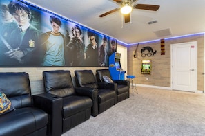 A home theater room with a large wall-mounted screen, black leather recliners, a ceiling fan, and a door with a sign.