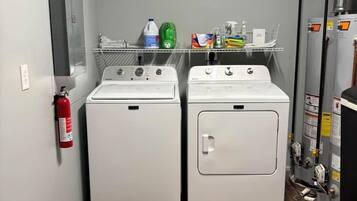 A laundry room with two front-loading washing machines, a fire extinguisher, and a shelf with cleaning supplies.