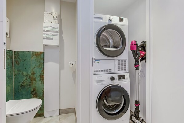 Une buanderie avec des machines à laver et des sèche-linge superposés, des toilettes et un mur rouillé.
