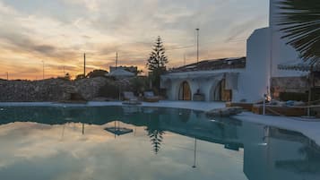 A pool with clear water reflecting trees and a sunset sky. There are lounge chairs and a stone wall in the background.