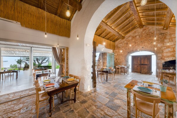 A spacious dining area with stone flooring, wooden tables, and chairs, featuring a large arched doorway and a view of the outdoors. - Masseria Samenti (Racale)