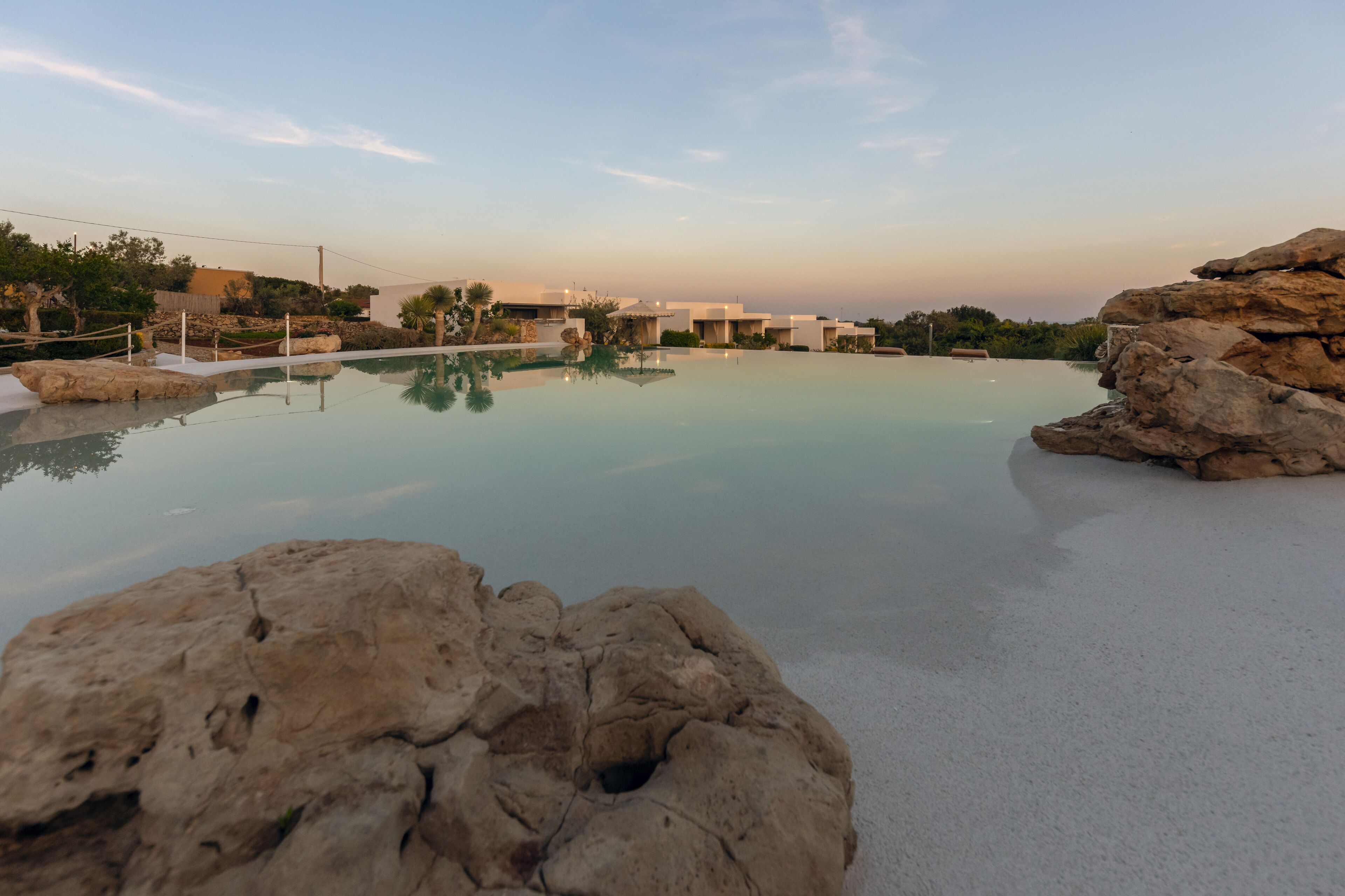 A resort pool with clear water, surrounded by rocks and palm trees, with buildings in the background.