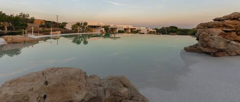 A resort pool with clear water, surrounded by rocks and palm trees, with buildings in the background.