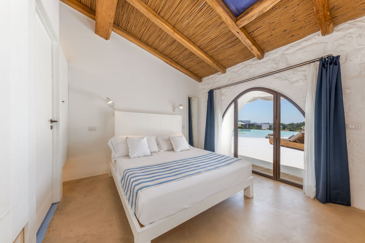 Une chambre avec un lit, un plafond en bois, une grande fenêtre avec des rideaux et une vue sur une piscine.