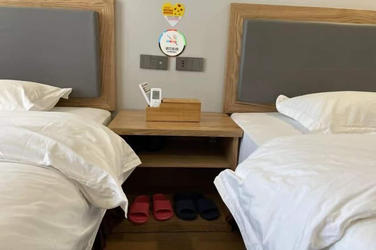 A hotel room with two beds, a nightstand with a phone and a small box, and a shoe rack with slippers.