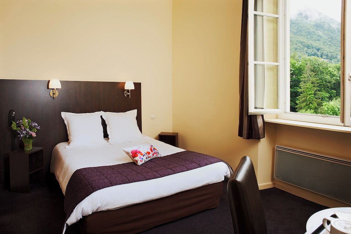 Une chambre d’hôtel avec un lit, une table de chevet ornée d’un vase de fleurs, une chaise, une table avec une tasse, et une fenêtre donnant sur un paysage verdoyant.