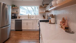 A modern kitchen with white cabinetry, a stainless steel refrigerator, a dishwasher, and a large window providing natural light. - As Featured on Staycation TV, A House, Cottage, & Tiny Home in Downtown Norcross (Norcross)