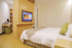 A hotel room with a bed, a desk with a computer, a television mounted on the wall, and a green trash bin.