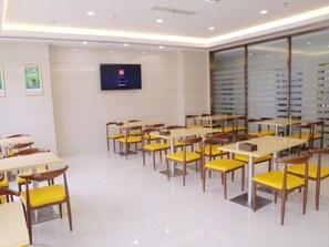 A modern dining area with yellow chairs and white tables, a flat-screen TV on the wall, and a large window with blinds. - Greentree Inn Hefei Feidong County High Speed Rail Station Wuyue Plaza Express Hotel (Hefei)