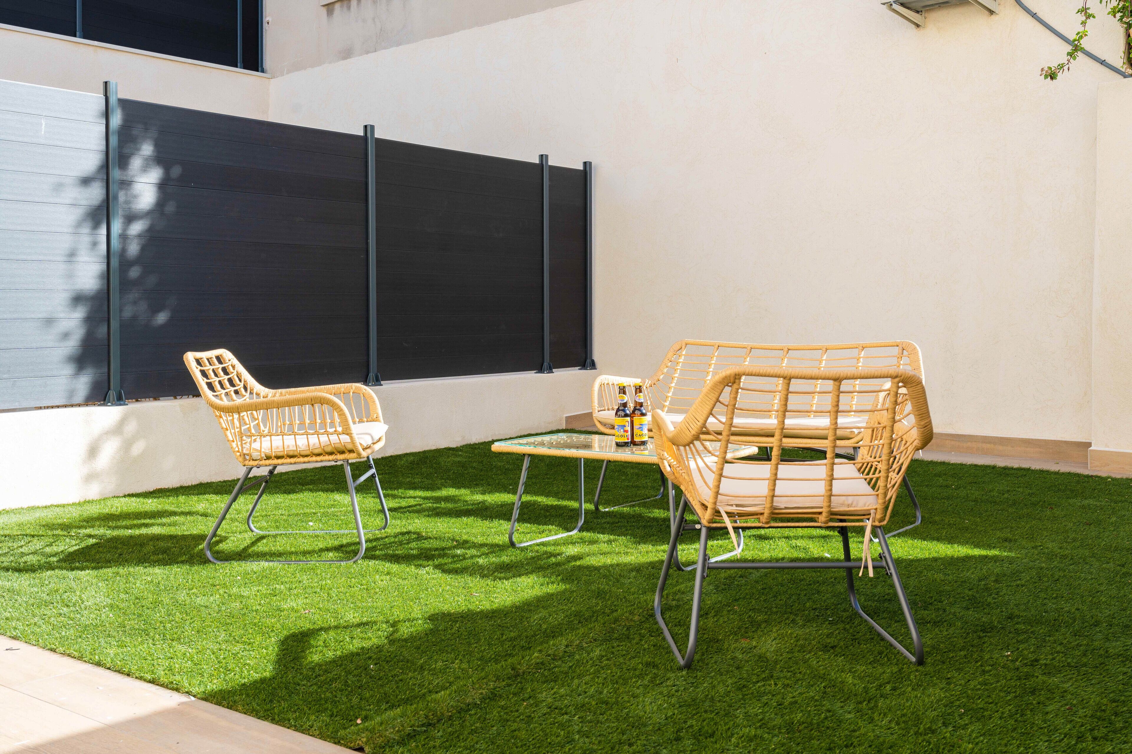 A patio with two wicker chairs and a small table, a black privacy screen, and artificial green grass.