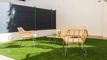 A patio with two wicker chairs and a small table, a black privacy screen, and artificial green grass.